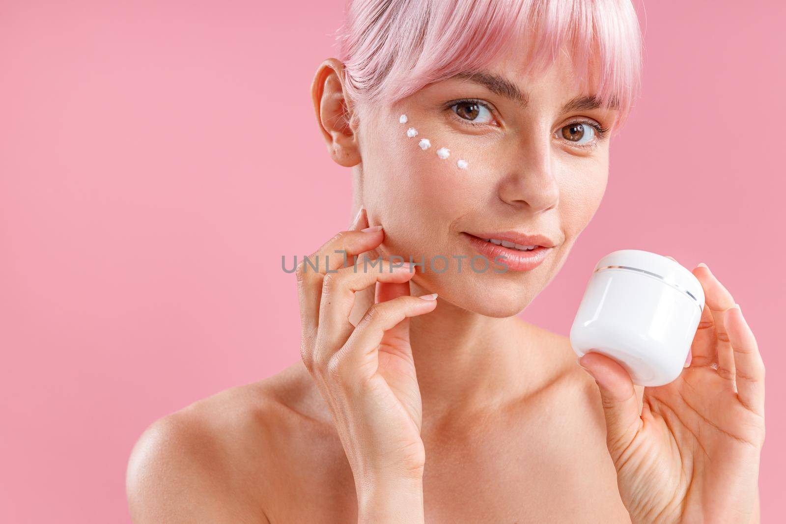 Portrait of beautiful woman with pink hair and face cream applied on her skin like dots looking at camera, holding cream in white jar isolated over pink background. Beauty, spa, skin care concept