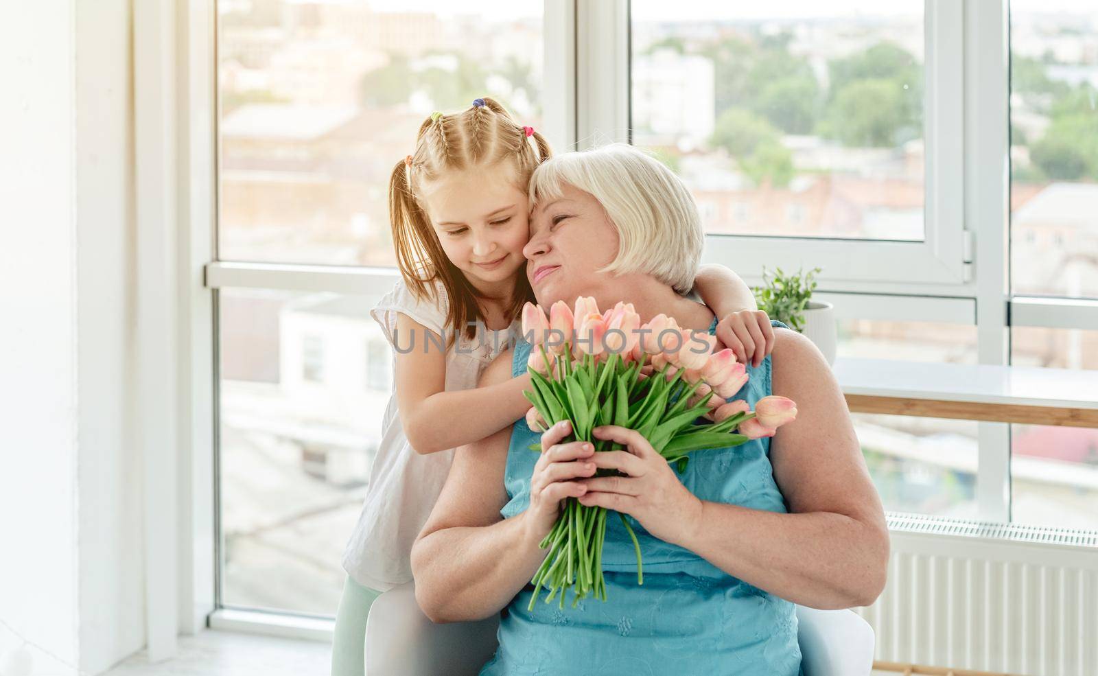 Pretty little girl kissing happy grandmother with bouquet of tulips indoors