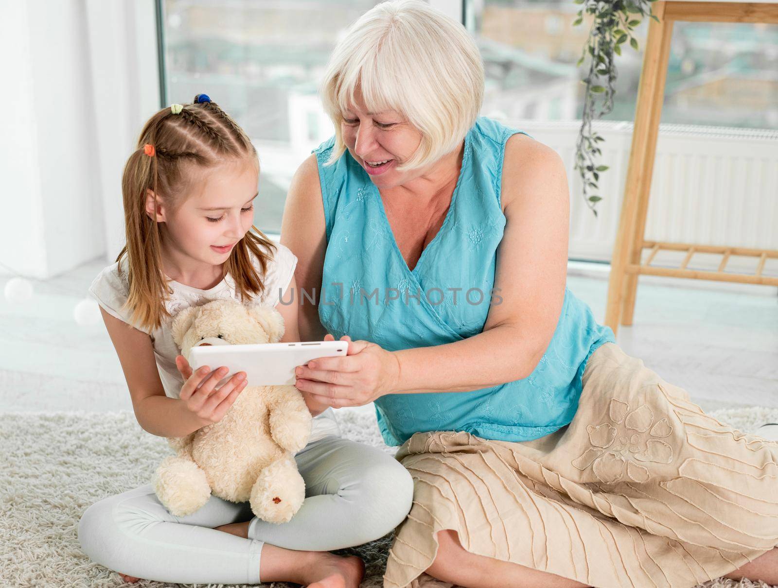 Happy grandmother with little granddaughter using tablet siting on floor in children's room