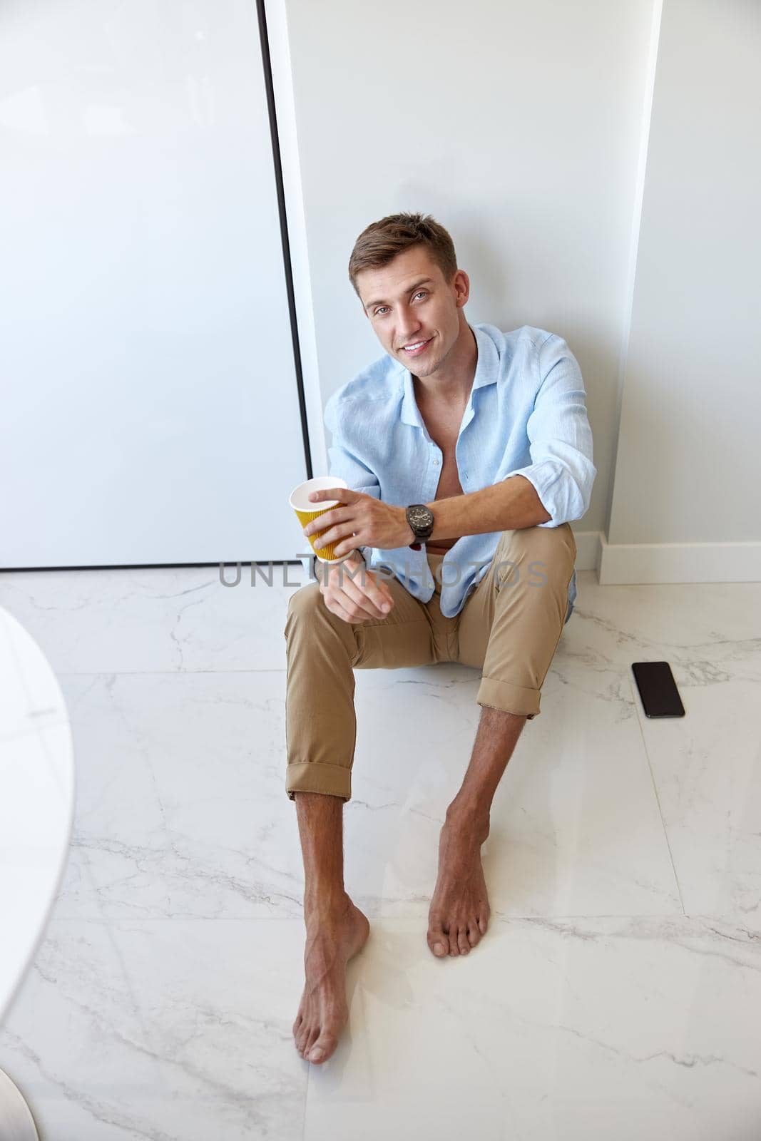 Caucasian hadsome young man in blue shirt is sitting on the floor and chilling at modern kitchen