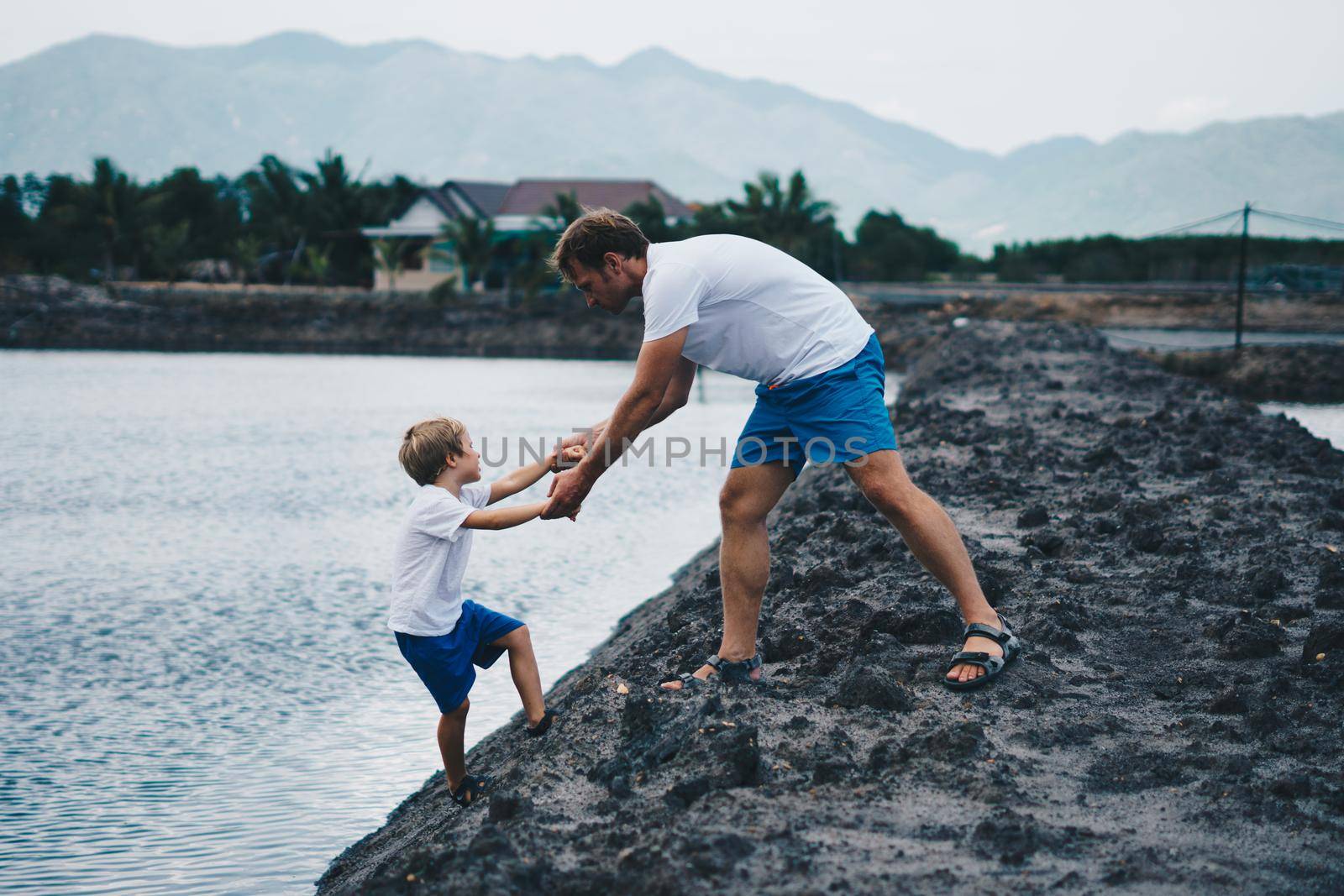 Dad helps son go down to lake water, explains safety rules. Evening, mountain background. Happy childhood fatherhood. Family together walk play. Home natural education, father's day, responsibilities.