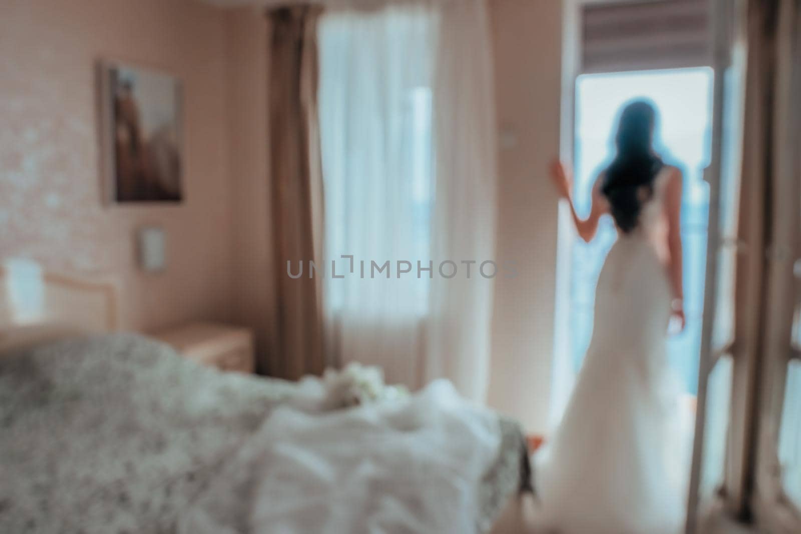 Blurred image for the background. Back view of beautiful brunette bride young woman in the white lace dress near window in the hotel room. Wedding concept. by panophotograph