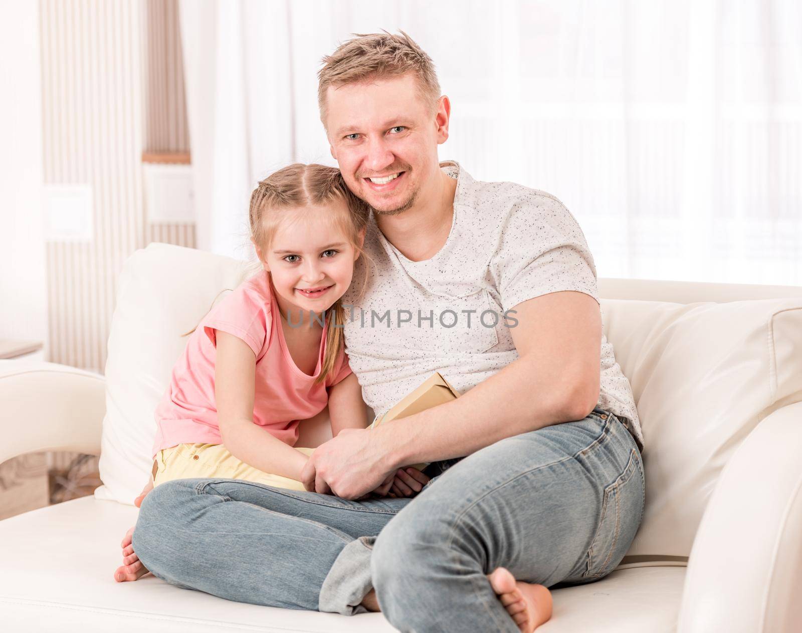 Handsome dad and cutie daughter sitting happy on the beige couch in guest room, with a fathers day gift