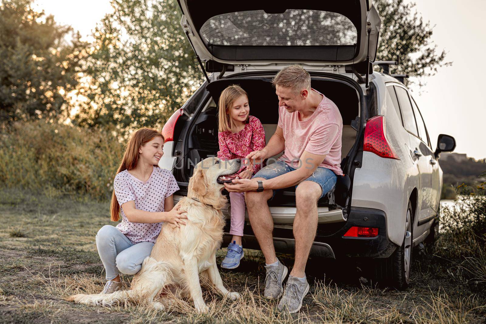 Father with daughters and golden retriever sitting in car trunk on nature