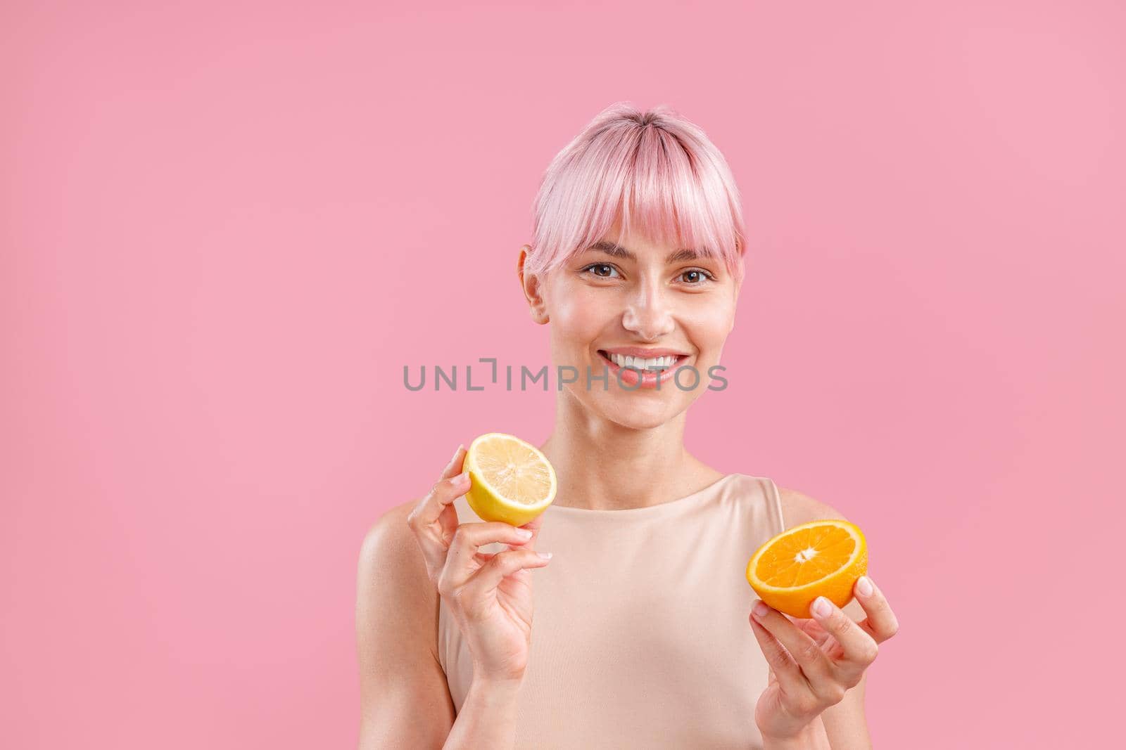 Portrait of smiling woman with pink hair holding half of fresh orange and lemon, posing isolated over pink background. Beauty, healthy eating concept
