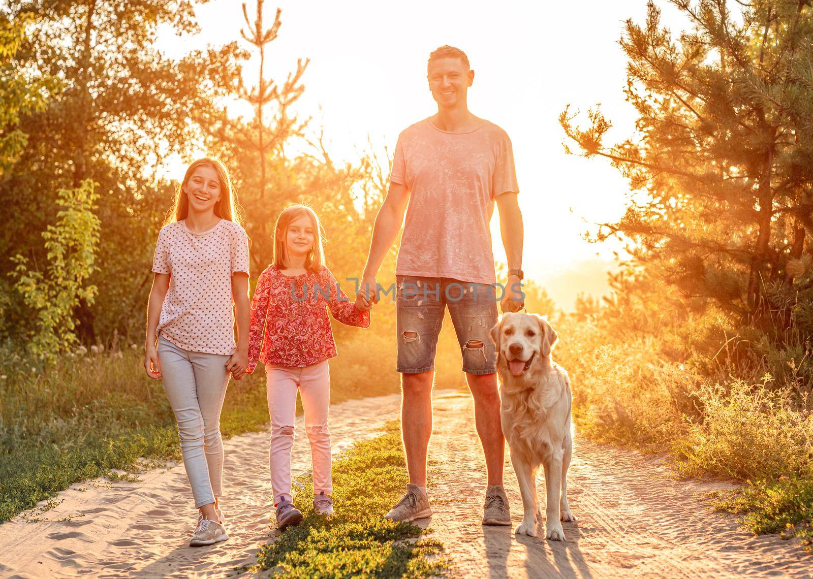 Father with daughters standing holding hands and golden retriever on leash outdoors at sunset