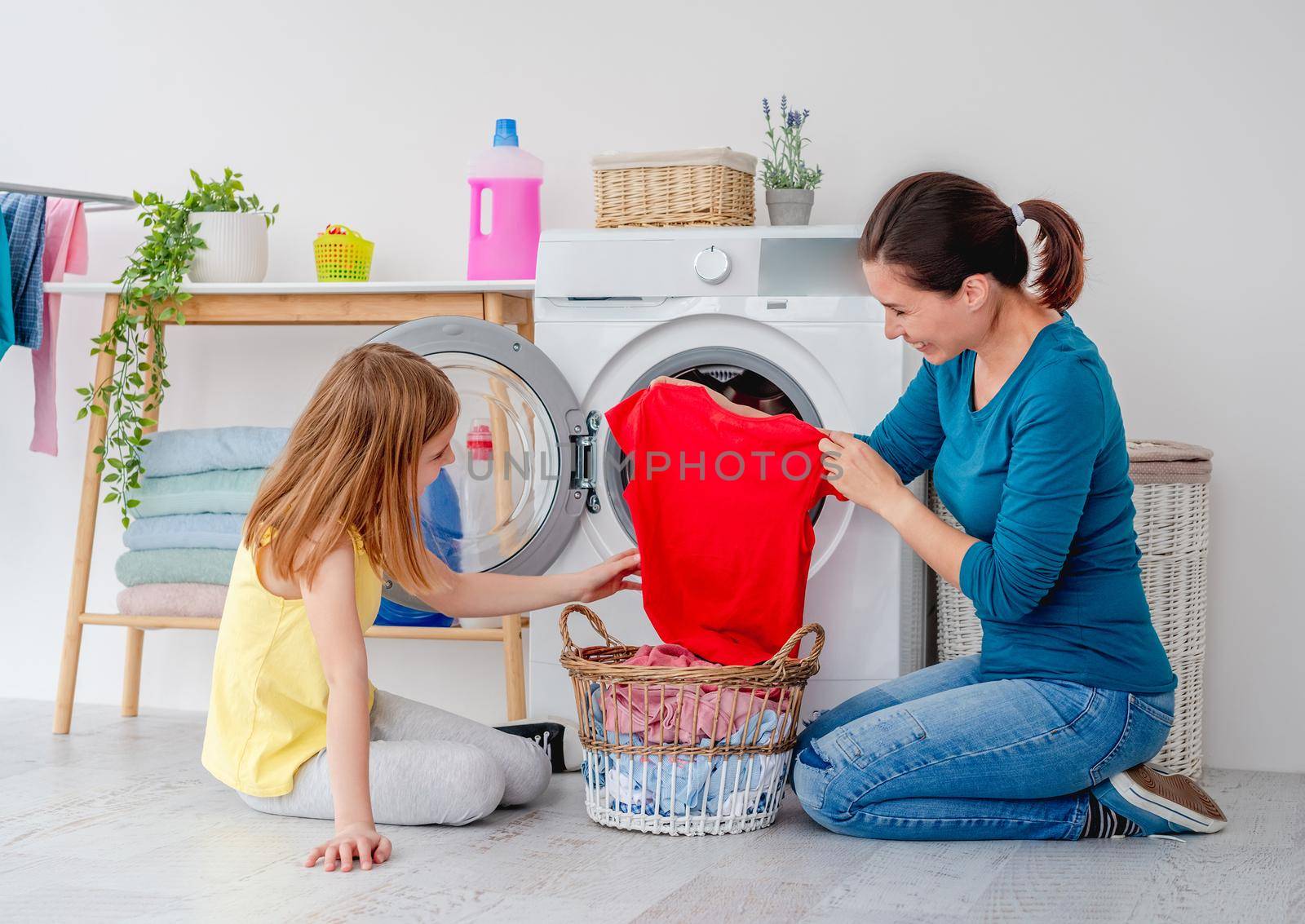 Mother with daughter sitting on floor near washing machine with clothes in basket in light bathroom