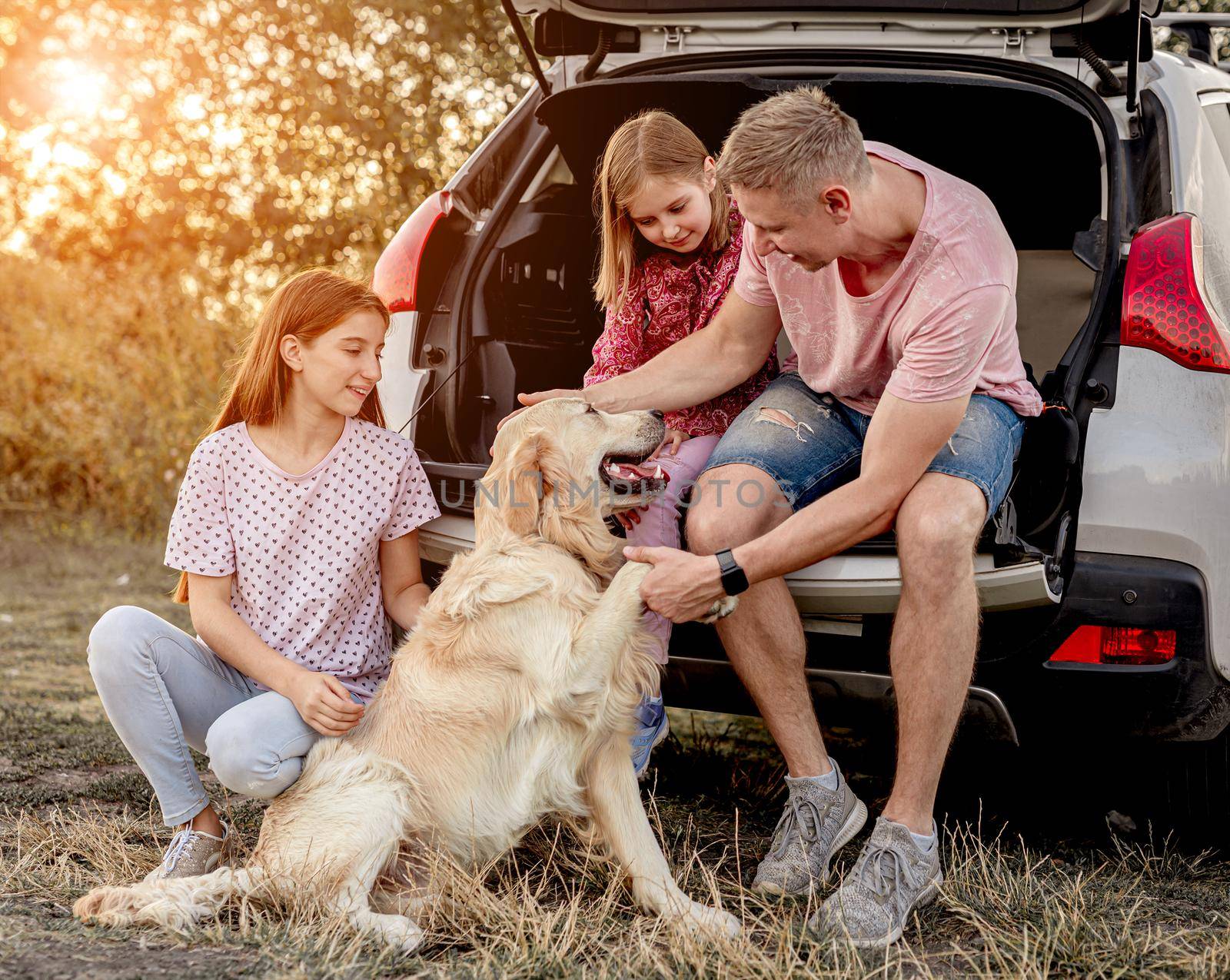 Family with golden retriever dog next to open car trunk on nature at sunset