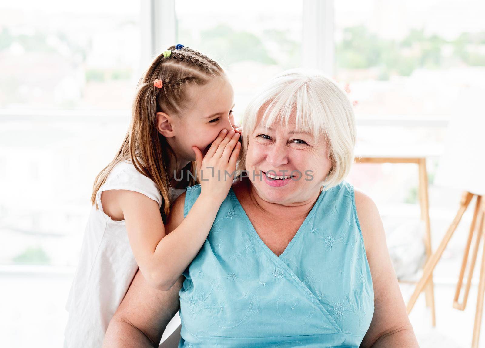 Happy little girl whispering secret to smiling grandmother in bright room