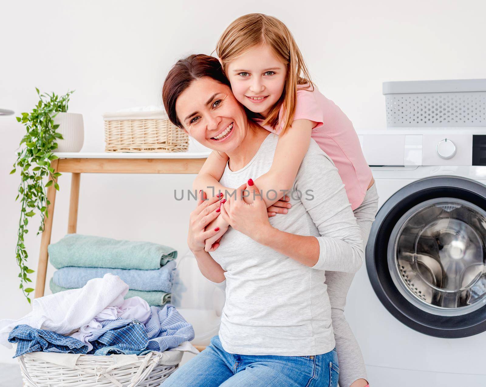 Little girl hugging with mother during laundry of clothes in washing machine at home