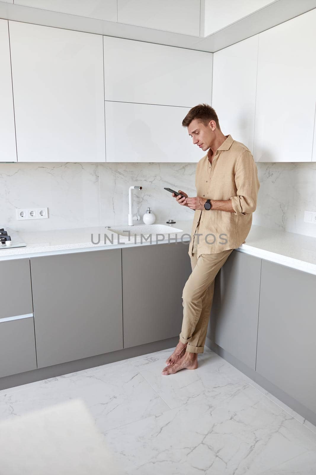 Hadsome smiling man is chilling on the minimalistic kitchen with smartphone by Yaroslav_astakhov