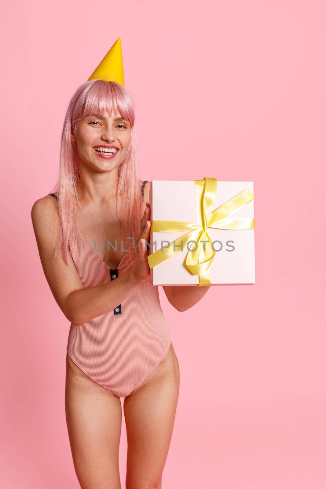 Smiling young woman with pink hair wearing swimsuit and party hat holding cute gift box, standing isolated over pink background. Holiday, birthday concept