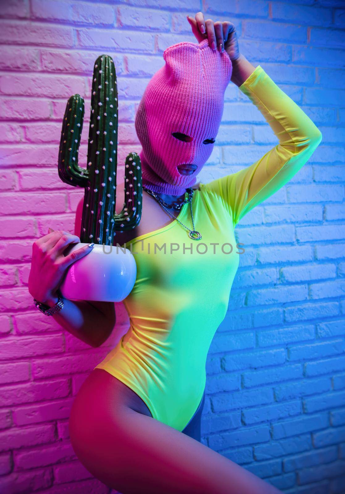 sexy woman in pink mask with cactus on white brick wall background in neon light by Rotozey