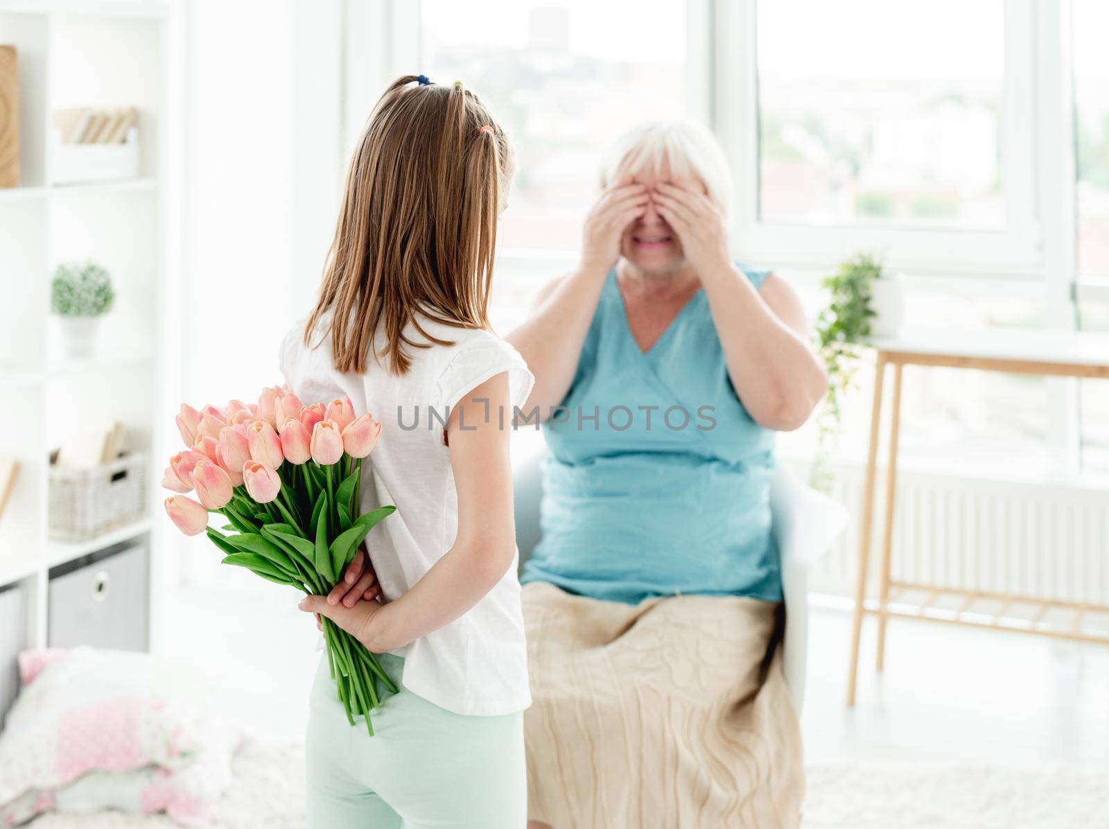 Rear view of little girl giving tulips to grandmother in light room