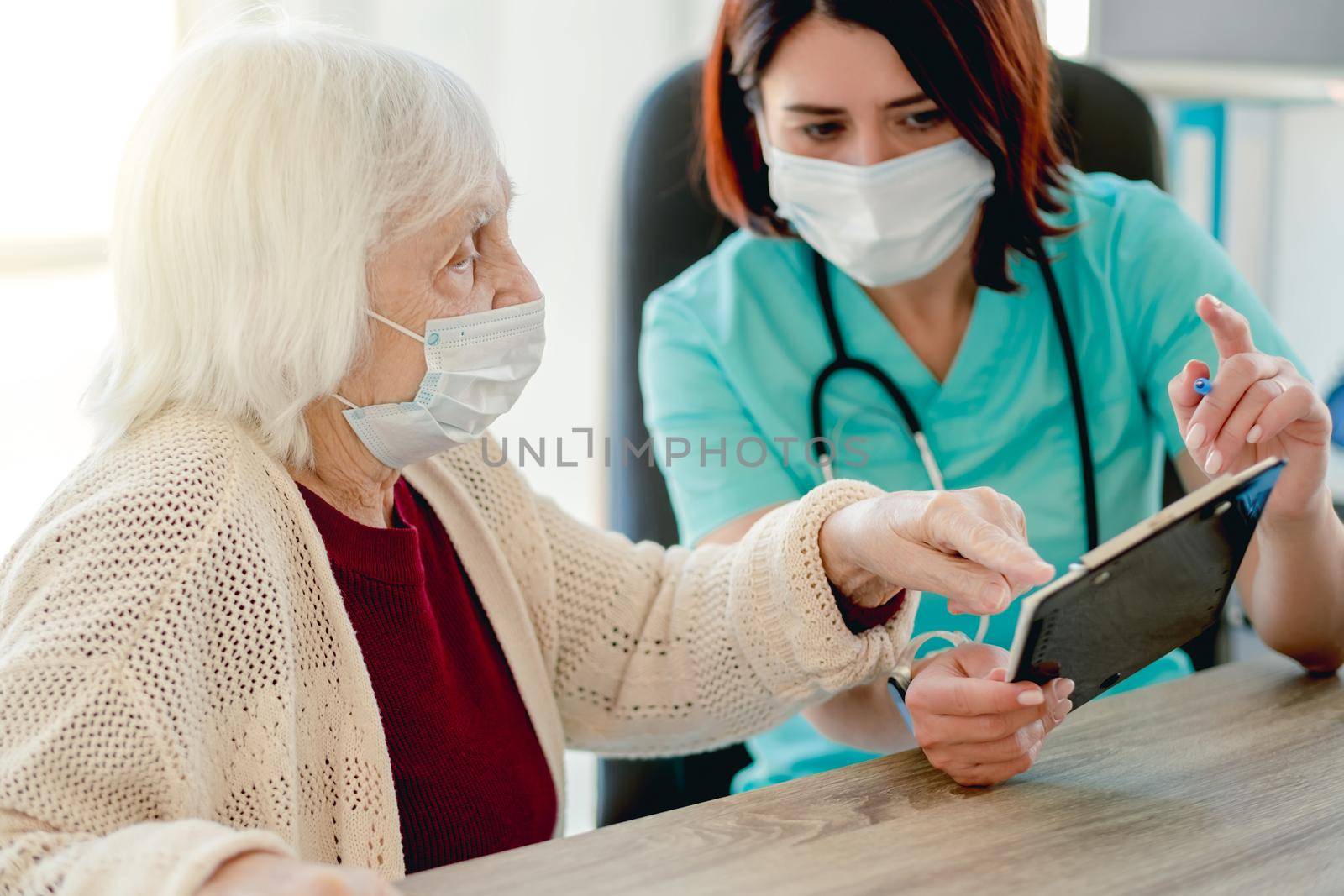 Therapist talking with old woman patient while holding papers and pen during appointment in clinic