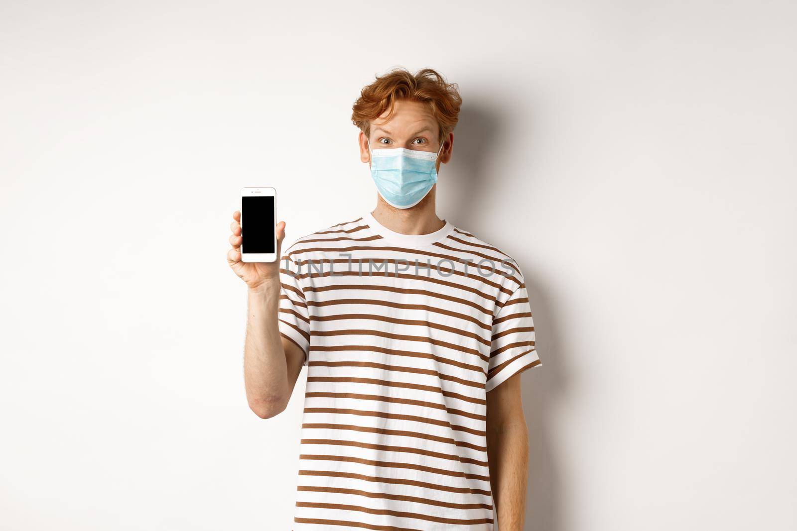 Covid-19, online doctor concept. Smiling redhead man in face mask showing smartphone screen, standing over white background.