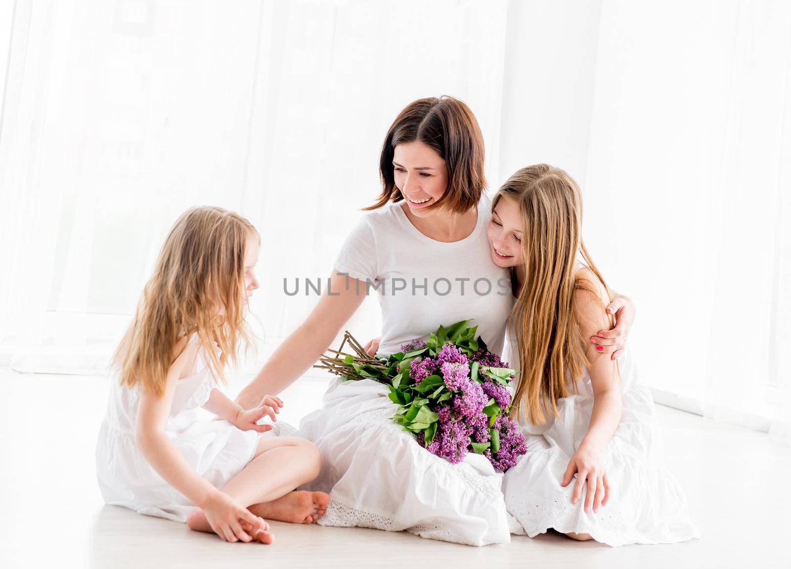 Young beautiful woman with her little daughters hugging and smiling while sitting on the floor with lilac bouquet. Mother with little daughters in white dresses in white light room