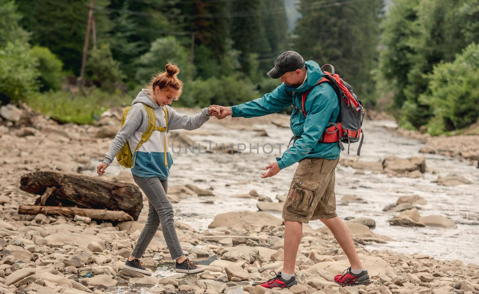 Man helping young girl to cross rocky mountain river during hiking together on spring nature