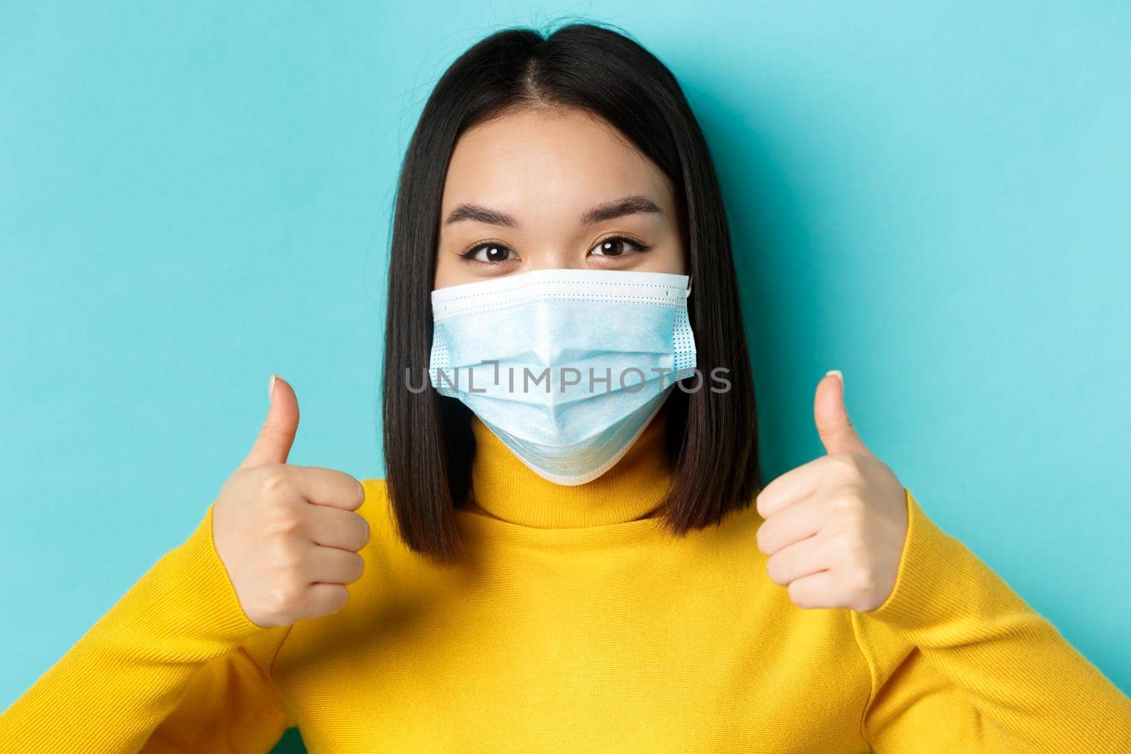 Covid-19, social distancing and pandemic concept. Close up of young asian woman in medical mask showing thumbs up, give approval, praise good offer, standing over blue background.