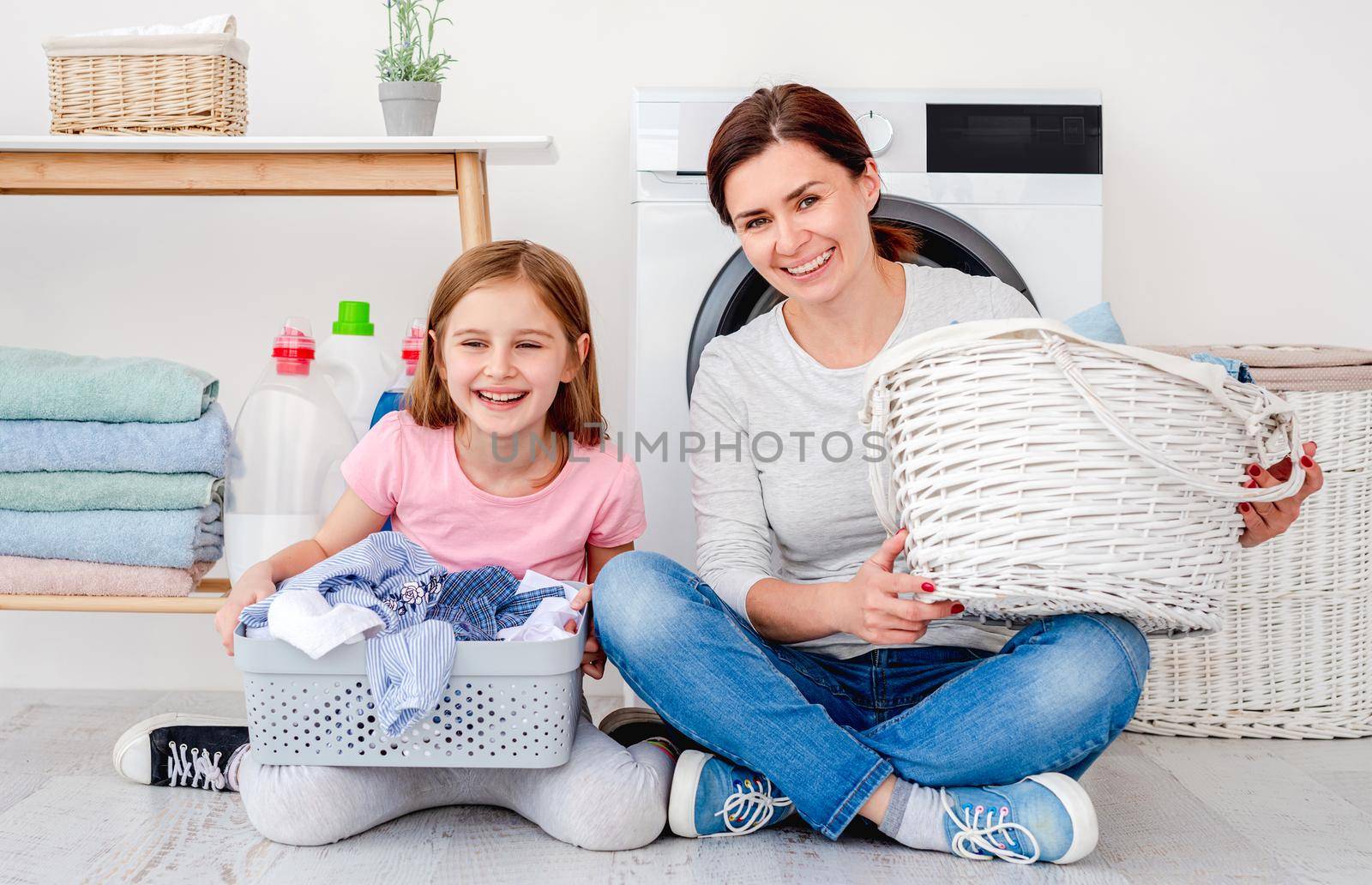 Mother and daughter having fun during laundry sitting on floor with washing baskets in light room