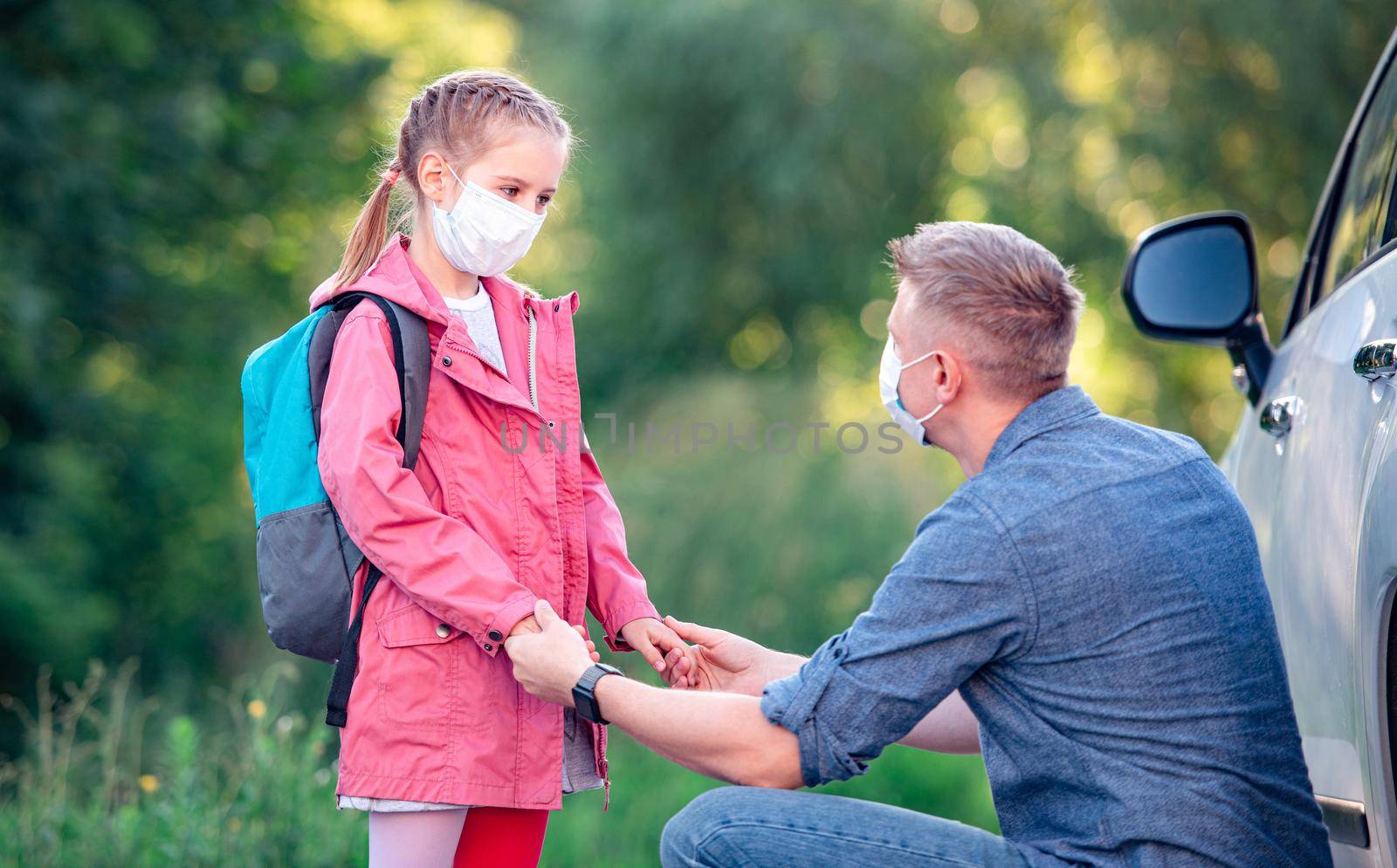 School girl with father in masks meeting after lessons outdoors near car
