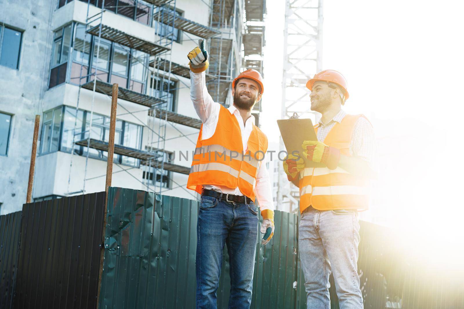 Two young male engineers in uniform and hardhats working at construction site, close up