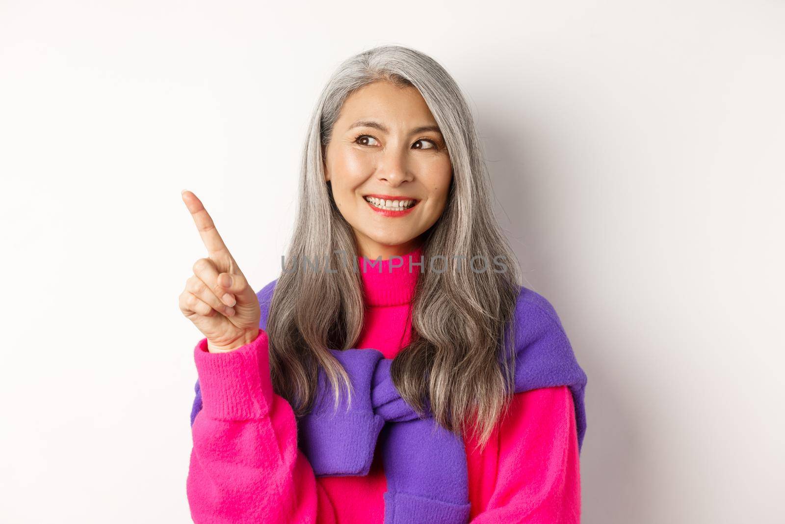 Close-up of beautiful asian senior woman smiling, looking and pointing upper left corner advertisement, checking out promo offer, standing over white background.