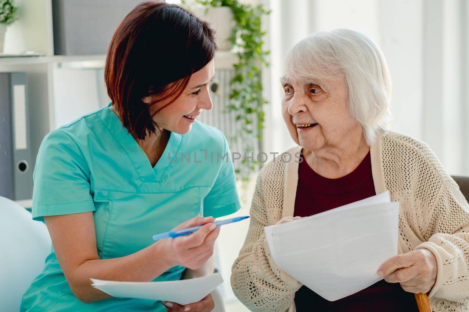 Therapist talking with old woman patient while holding papers and pen during appointment in clinic
