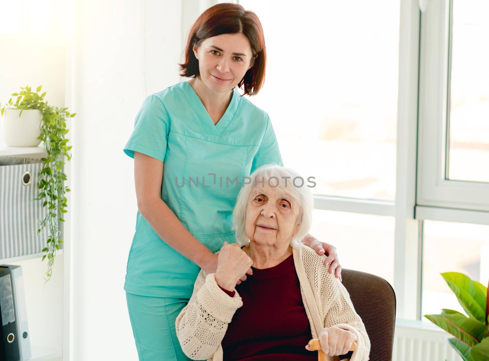 Caregiver standing beside elderly woman on chair in light room inside nursing home