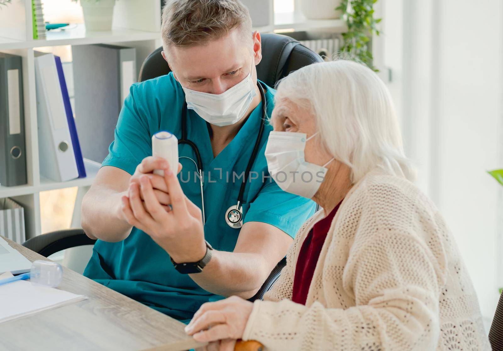 Therapist showing digital contactless thermometer to old woman at appointment during coronavirus pandemic