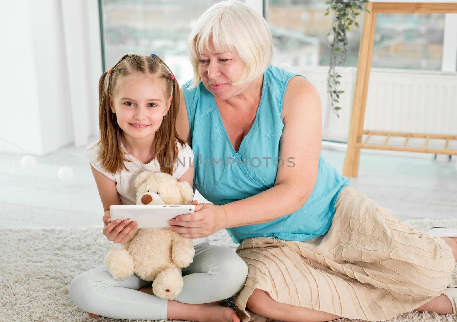 Happy grandmother with little granddaughter using tablet siting on floor in children's room