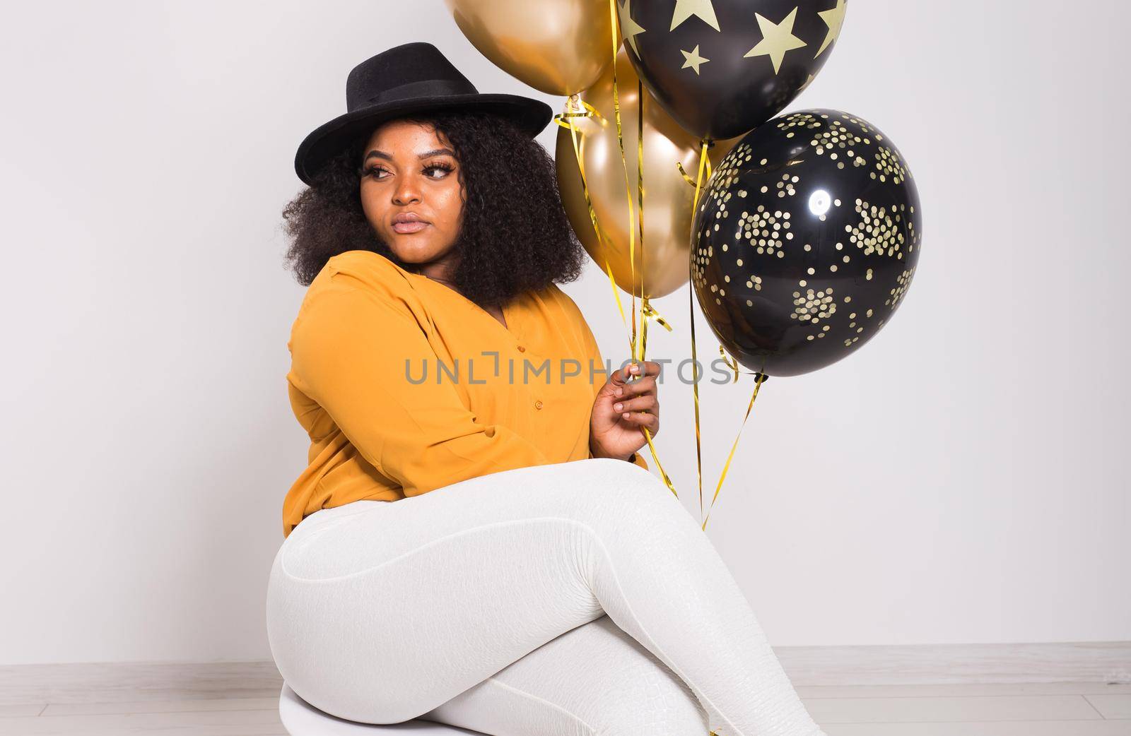 Portrait of smiling young African-American adult woman looking sweet on yellow background holding balloons.