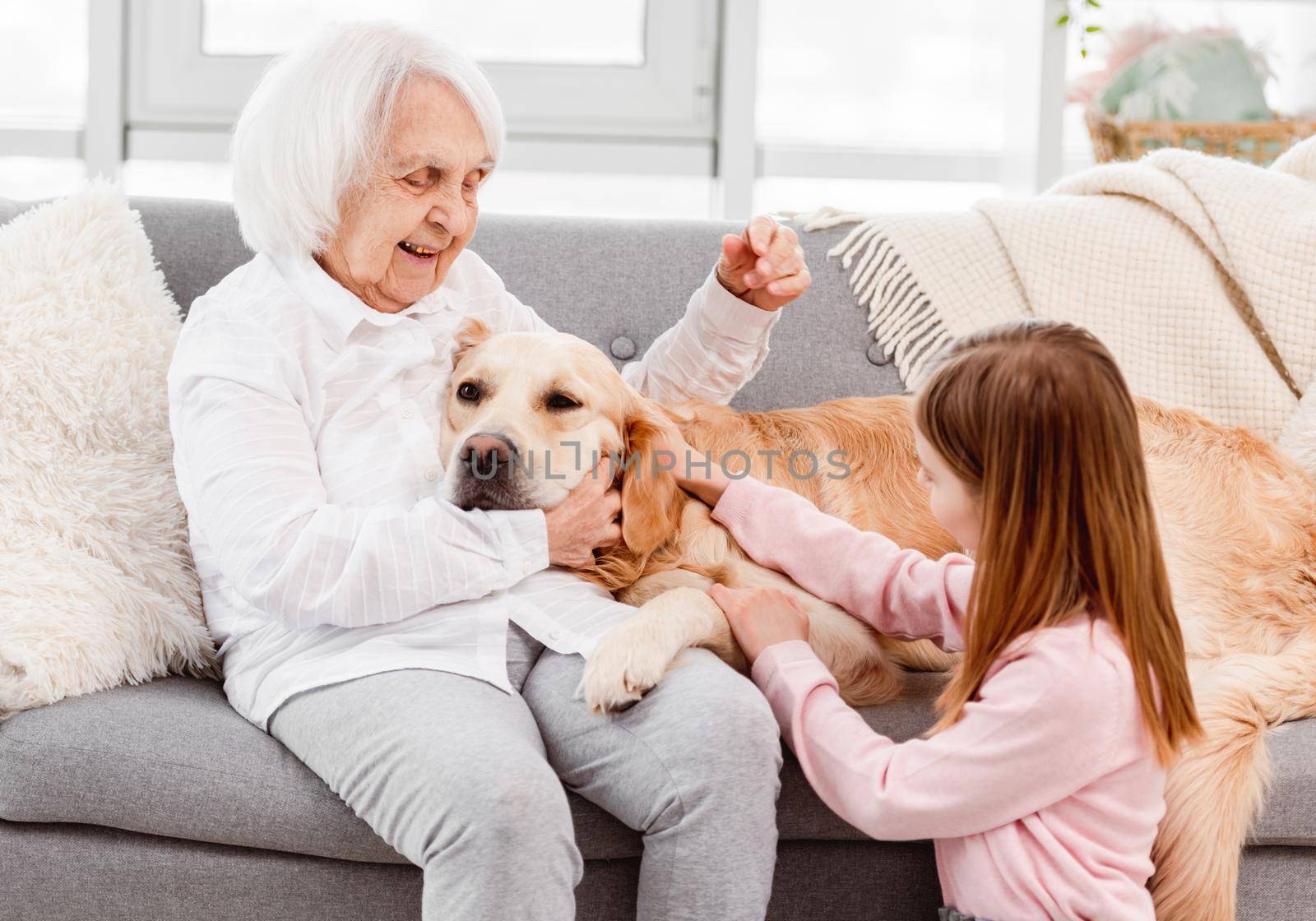 Grandmother with granddaughter hugging and petting golden retriever dog at home in sunny day