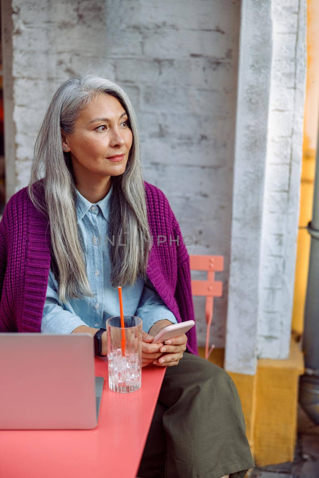 Beautiful mature Asian woman holds mobile phone sitting at coral table with notebook by Yaroslav_astakhov