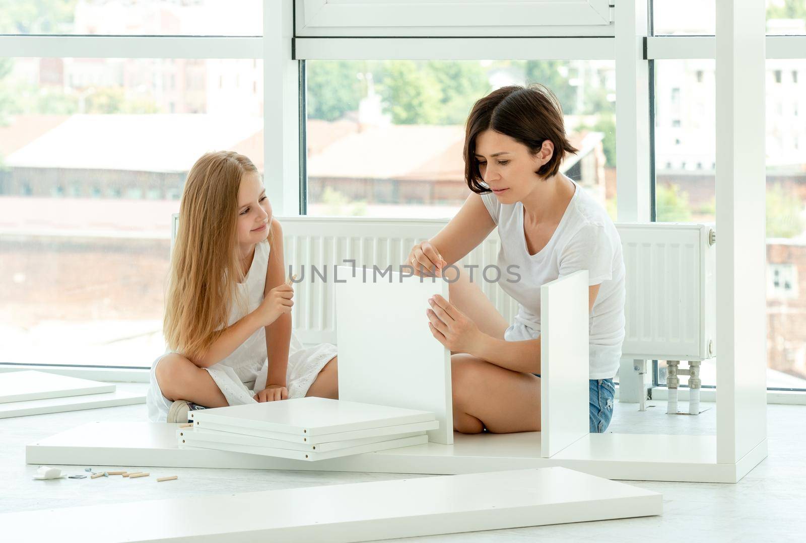 Lovely preteen girl helping her mother assembling new f completely white furniture at home together