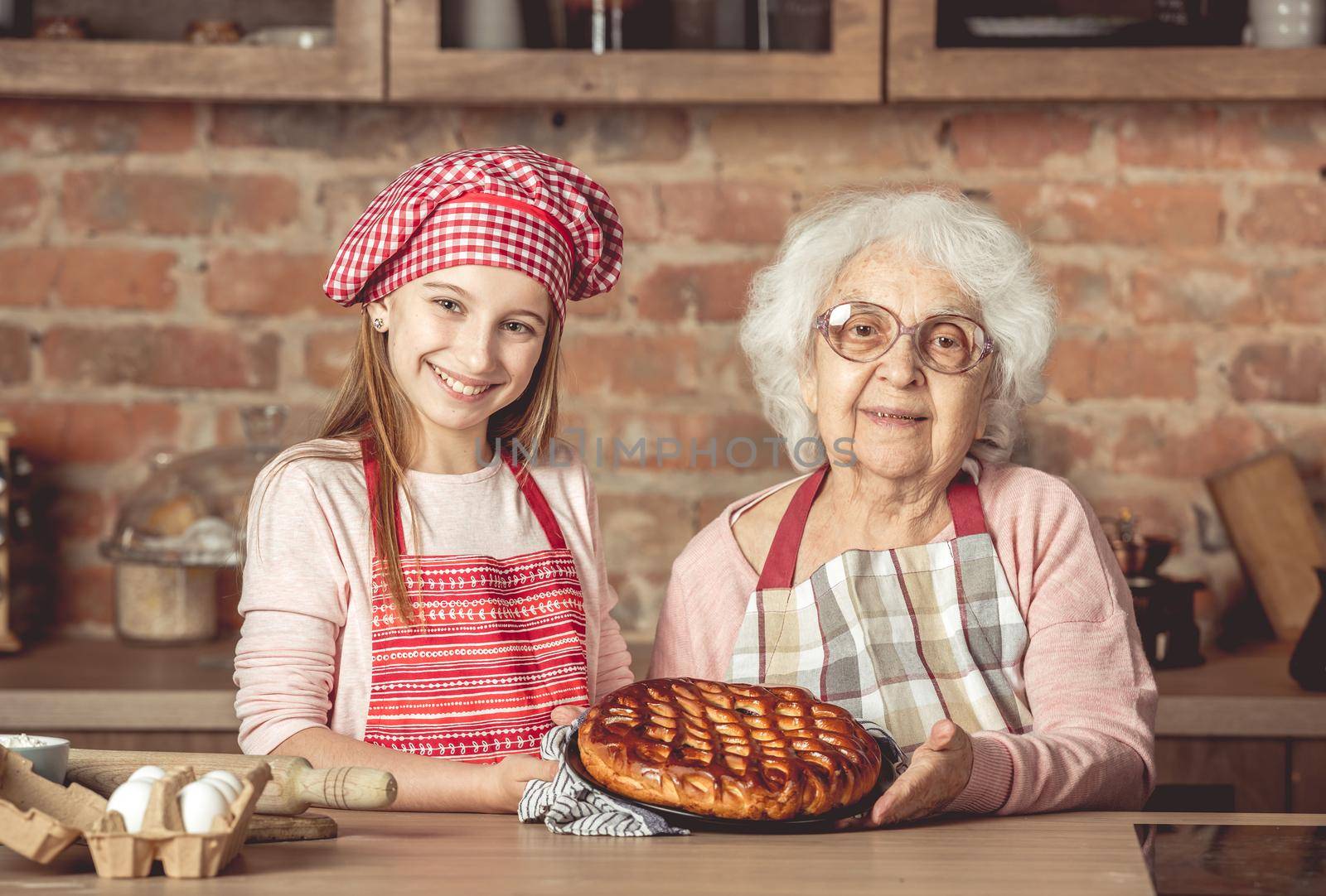 Young little granddaughter with her granny holding a tray and showing homemade fruit pie that they baked together