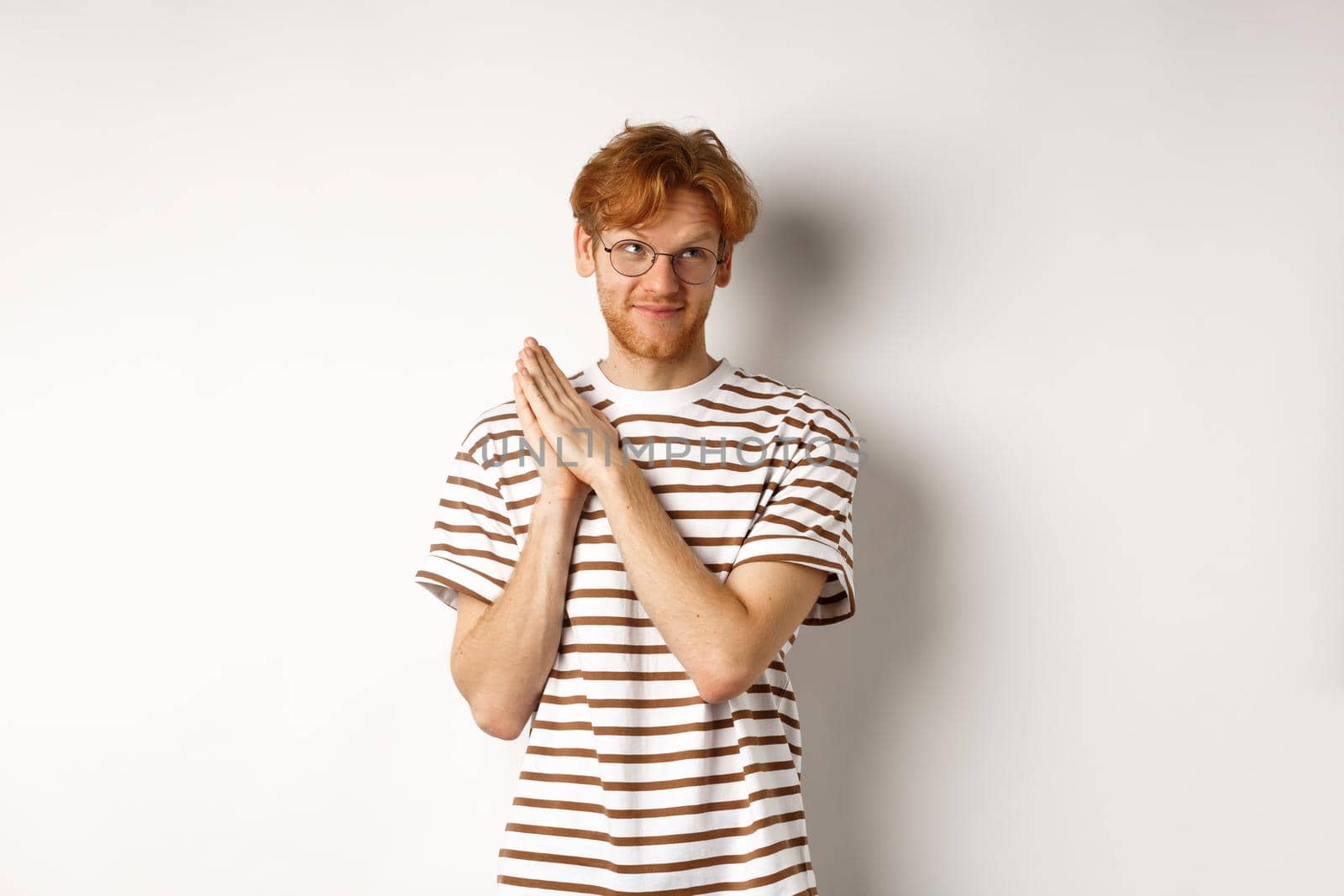 Thoughtful man with red hair rubbing hands and looking at upper right corner, having an idea, standing over white background.