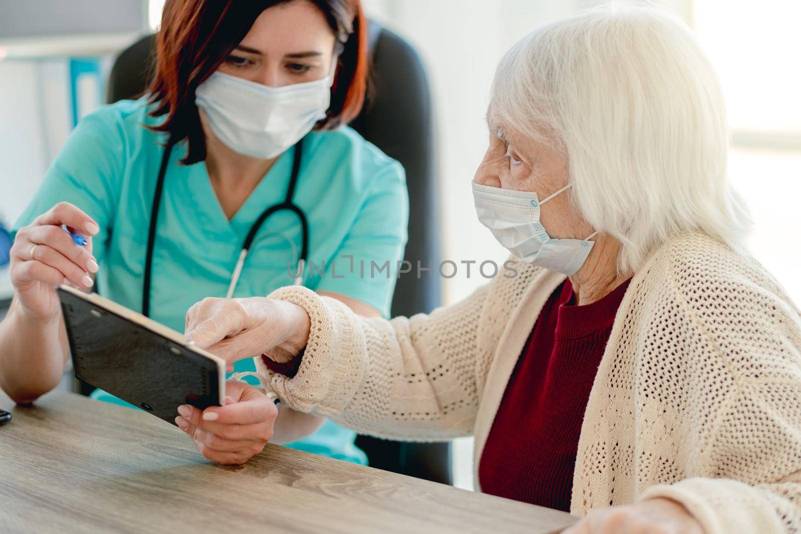 Therapist talking with old woman patient while holding papers and pen during appointment in clinic