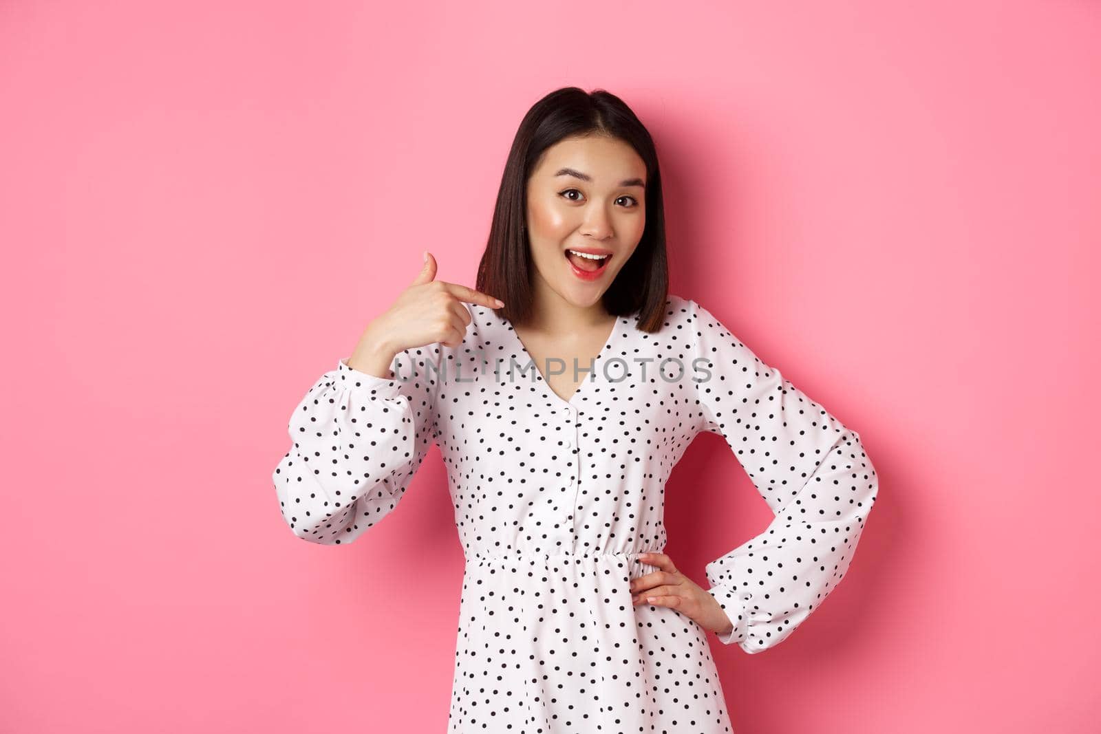 Beautiful asian girl in dress looking happy, pointing finger at herself, standing on romantic pink background.