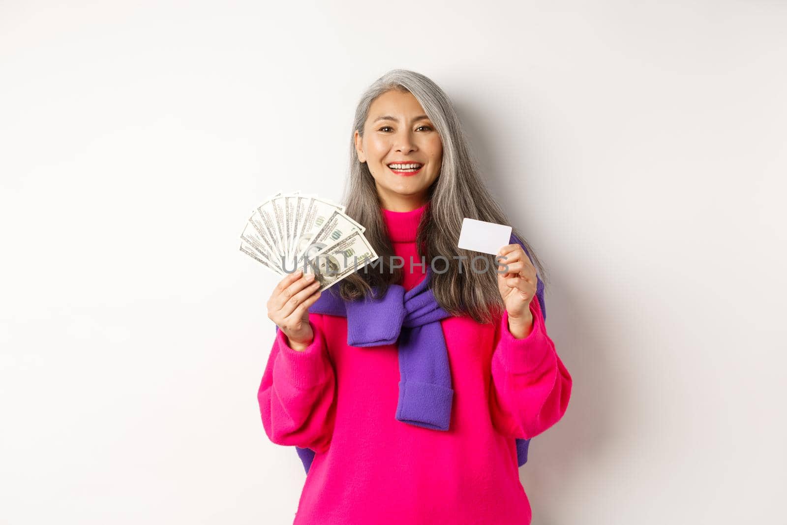 Shopping concept. Stylish asian senior woman with grey hair, showing money dollars and plastic credit card, standing over white background.