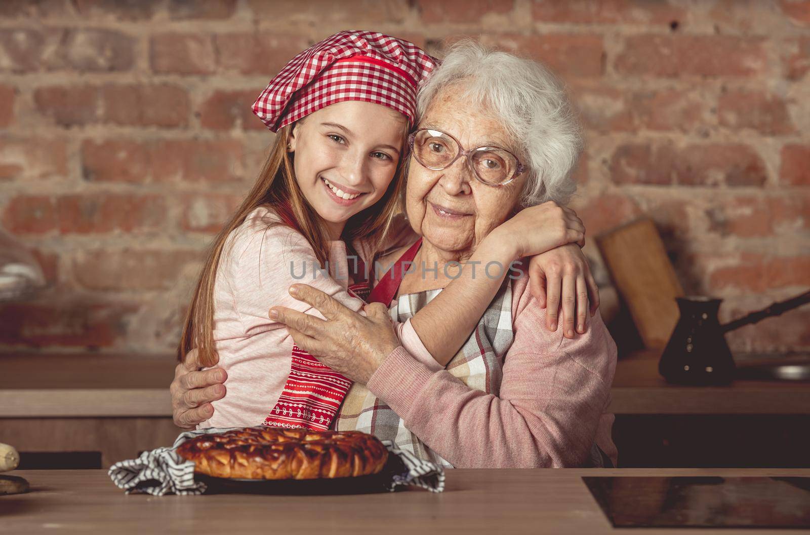 Little granddaughter hug her happy grandmother sitting at rustic kitchen at home