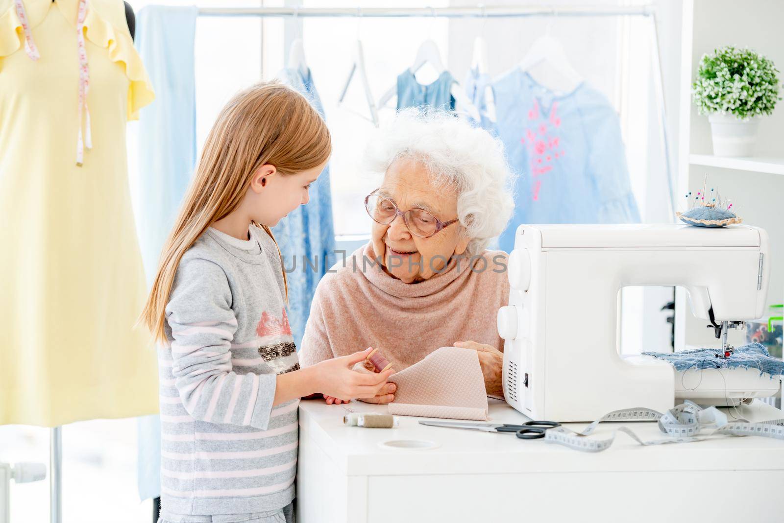 Elderly woman teaching little girl to sew in atelier