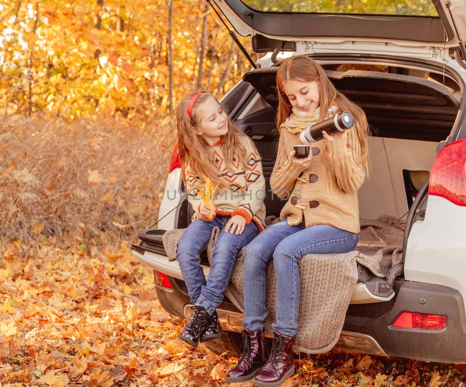 Pretty sisters sharing tea from thermos sitting in car trunk in autumn park