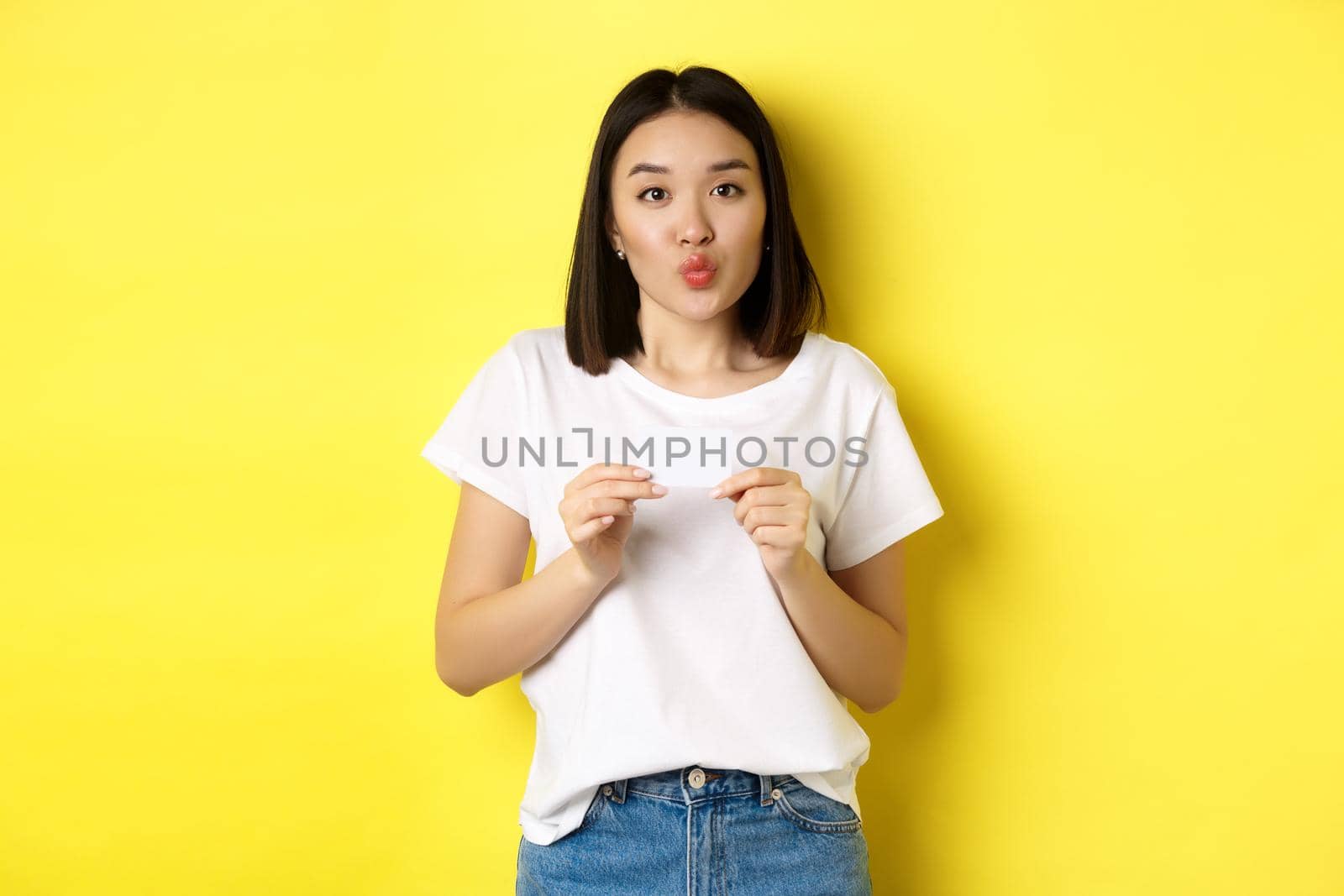 Young asian woman in casual white t-shirt showing plastic credit card and pucker lips silly, going on shopping, yellow background by Benzoix