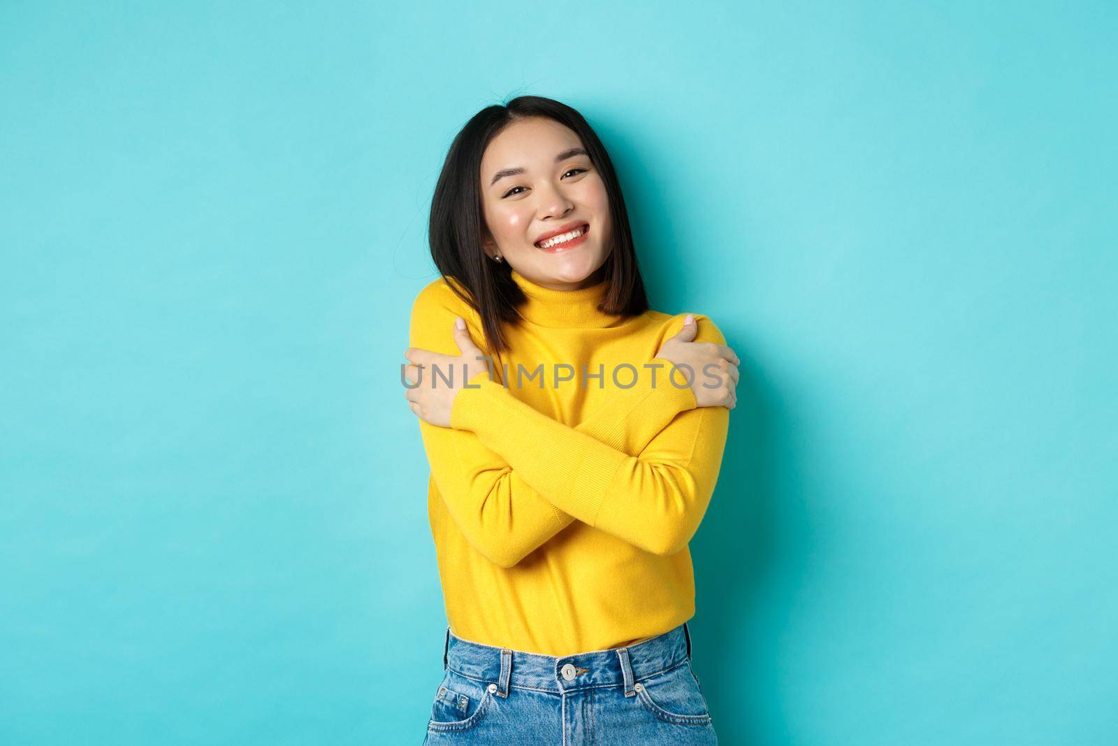 Cheerful beautiful girl embracing own body, smiling and cuddling herself, standing carefree in yellow pullover against blue background.