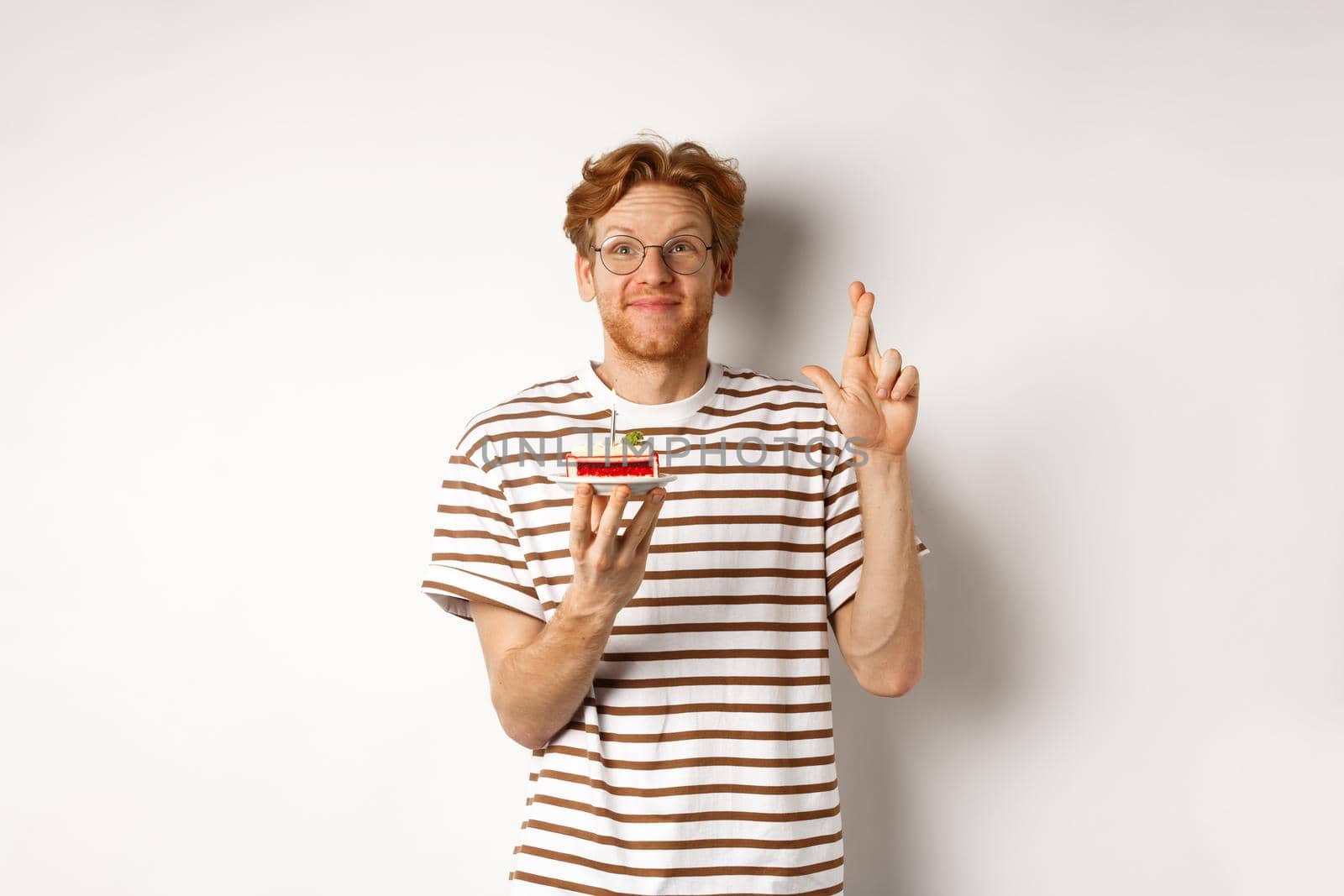 Holidays and celebration concept. Cheerful redhead man in glasses holding birthday cake with candle, cross fingers for good luck and making wish, white background.