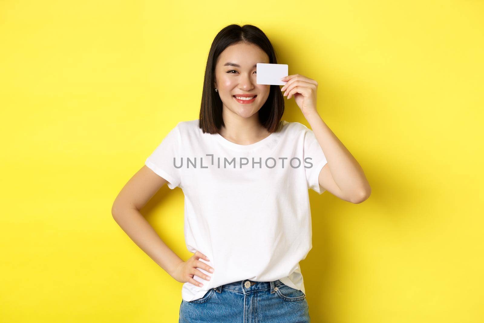 Young asian woman in casual white t-shirt showing plastic credit card and smiling at camera, yellow background by Benzoix