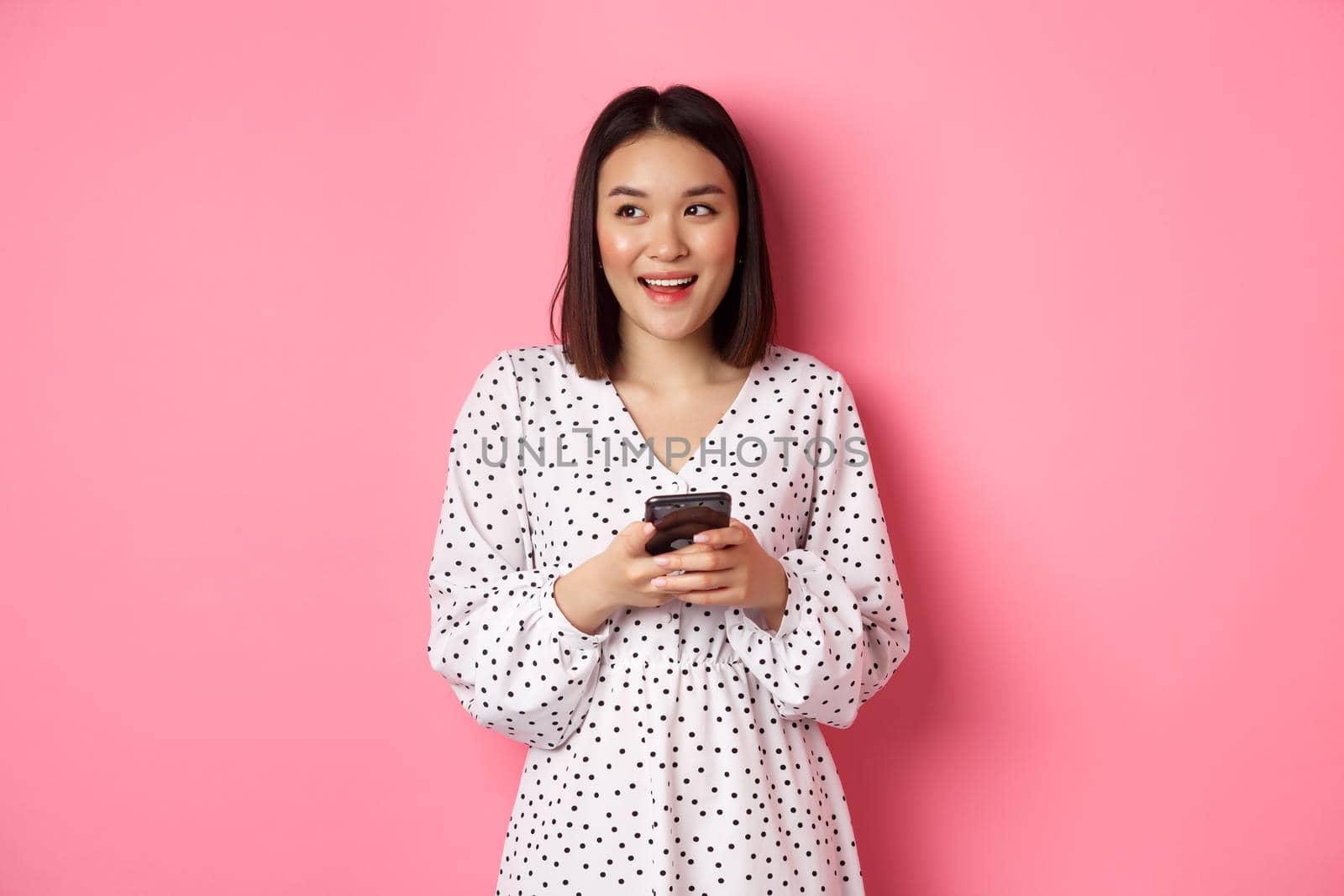 Cute asian woman thinking and smiling, looking left dreamy while messaging on smartphone, browing online stores, standing over pink background by Benzoix
