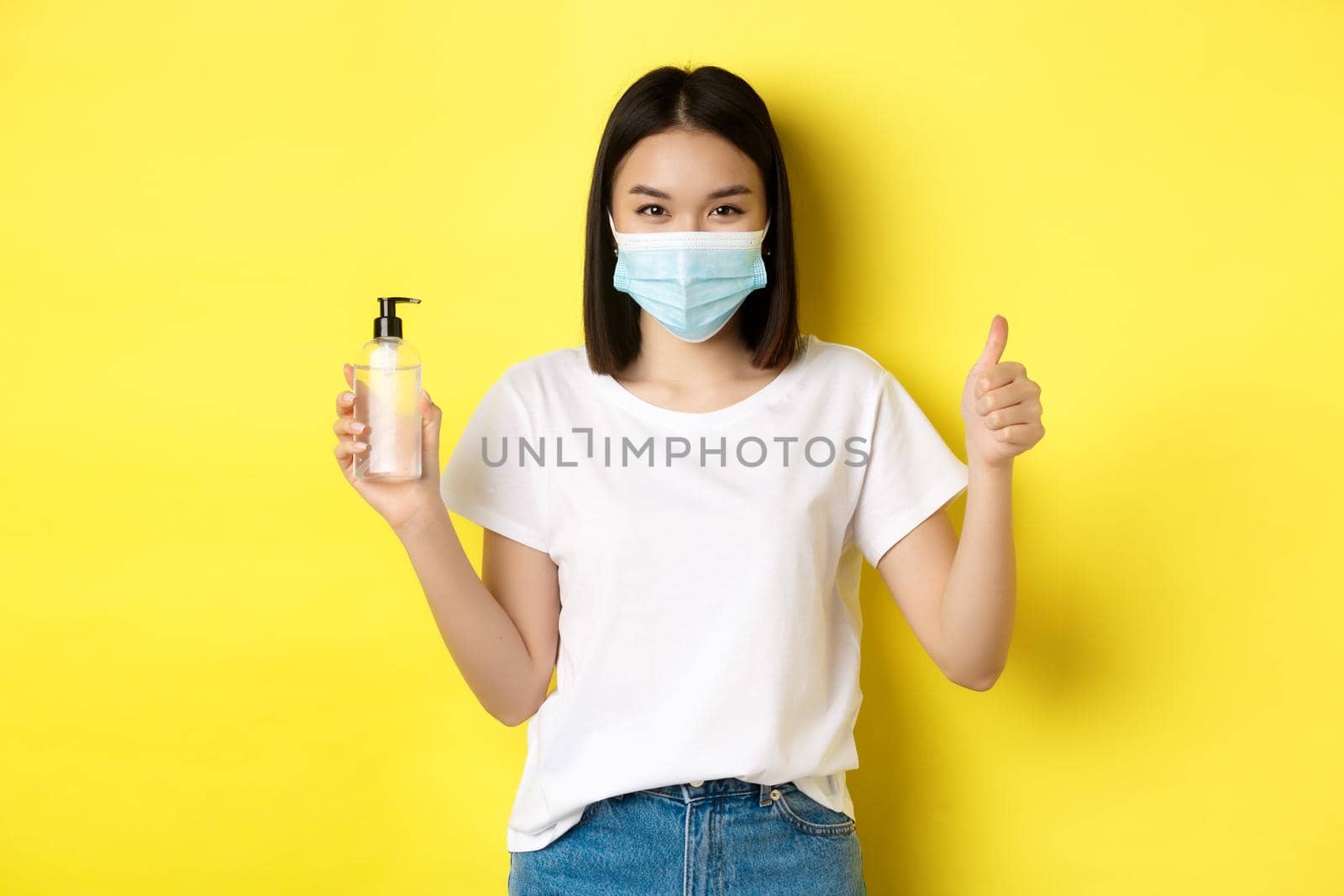 Covid, health care and pandemic concept. Asian girl in face mask from coronavirus, showing thumb up and good hand sanitizer, standing over yellow background by Benzoix