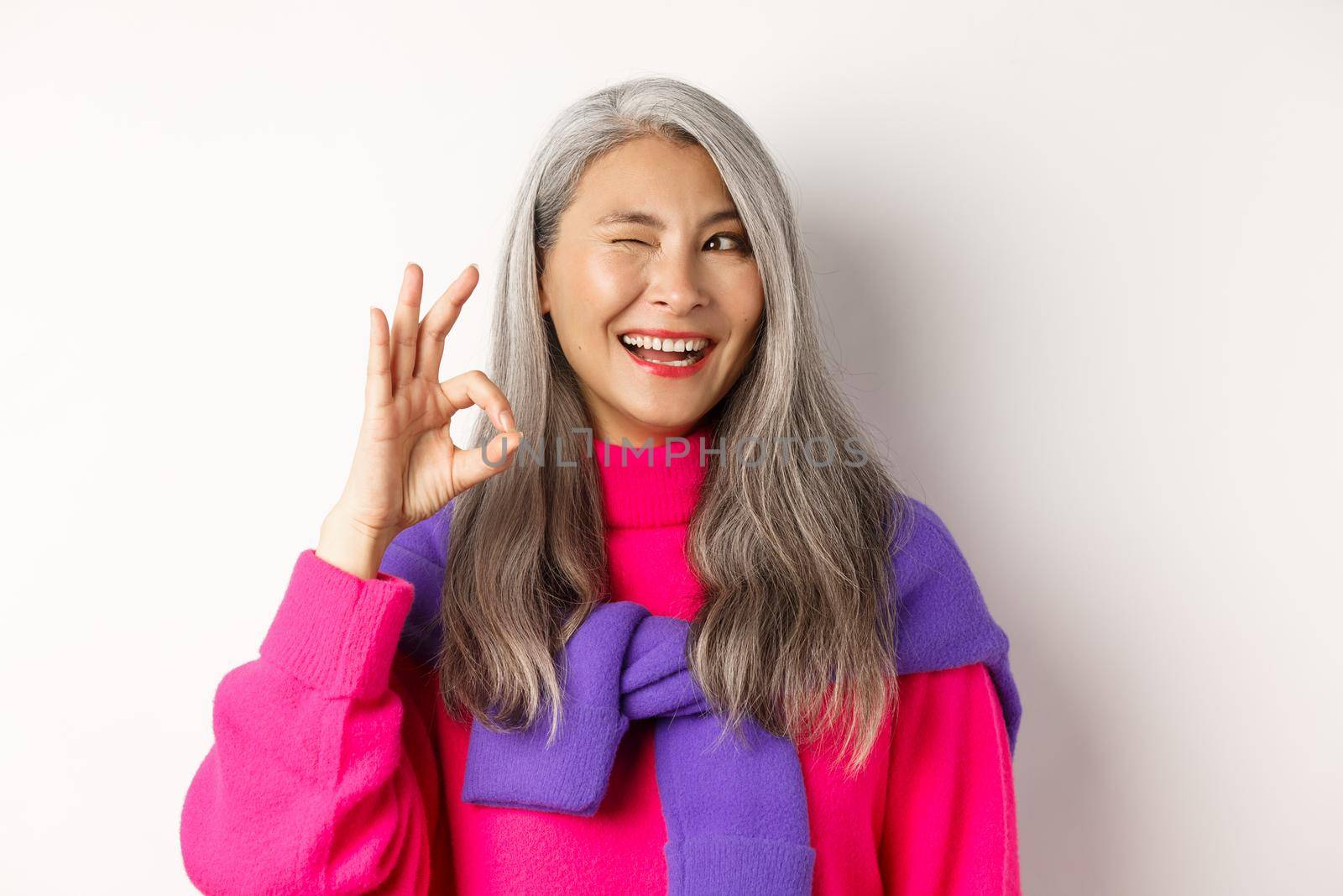 Close-up of cheerful asian grandmother in fashionable pink sweater, winking and showing okay sign, recommending promotion, standing over white background.